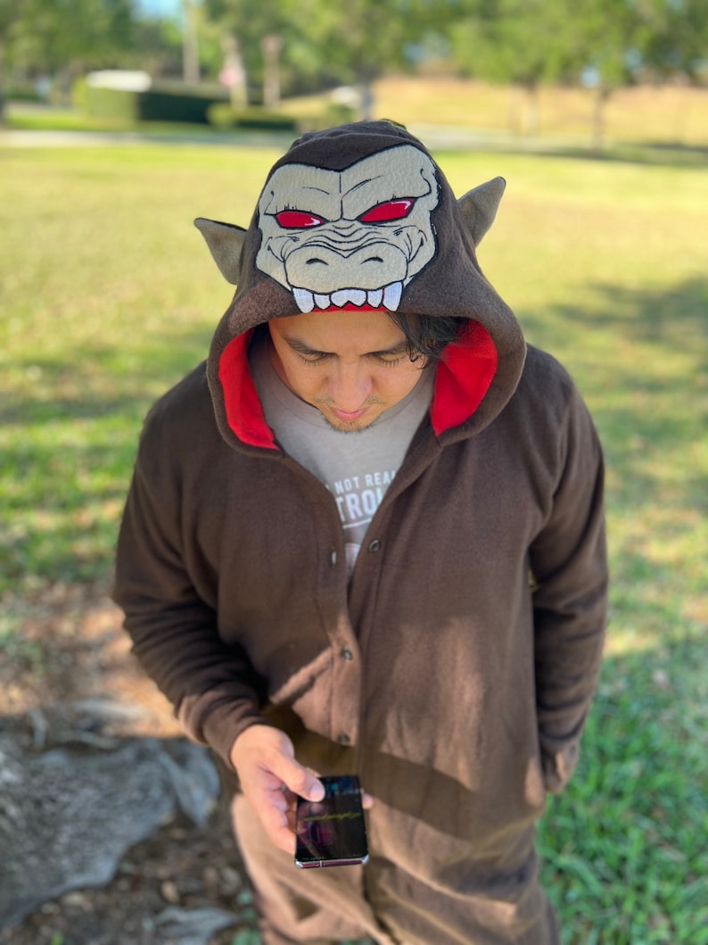 May include: A brown hooded jumpsuit with a red lining and a large embroidered monkey head on the hood. The monkey head has red eyes and sharp teeth. The person wearing the jumpsuit is looking down at a black smartphone.