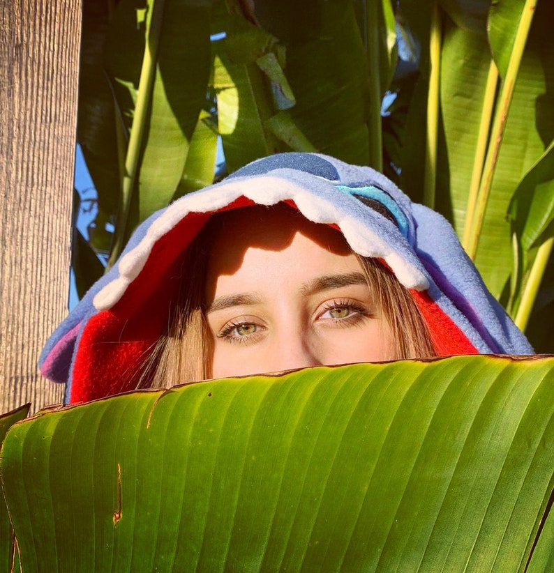 Puede incluir: Una persona lleva una sudadera con capucha azul y roja con un gran dise&ntilde;o de diente blanco. La persona est&aacute; parcialmente oculta detr&aacute;s de una gran hoja verde.