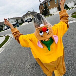 Puede incluir: Un disfraz de personaje de dibujos animados amarillo y marr&oacute;n con una capucha a juego. La capucha tiene una nariz roja, orejas rosas y una hoja verde. El disfraz se usa al aire libre en una carretera pavimentada.