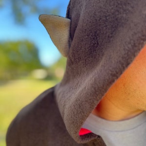 May include: A brown hooded sweatshirt with a light brown ear-like detail on the hood. The hood is pulled up over the head of the person wearing the sweatshirt. The sweatshirt has a red trim around the hood opening.