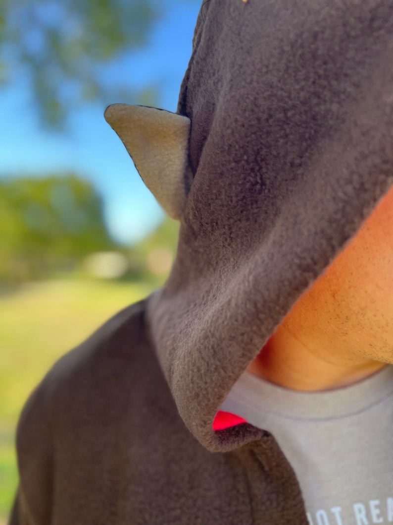 May include: A brown hooded sweatshirt with a light brown ear-like detail on the hood. The hood is pulled up over the head of the person wearing the sweatshirt. The sweatshirt has a red trim around the hood opening.