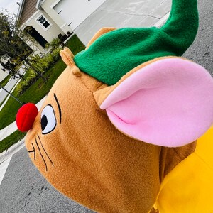 Puede incluir: Un disfraz de rat&oacute;n marr&oacute;n con un gorro verde, orejas rosas y una nariz roja. El disfraz tiene un dise&ntilde;o de dibujos animados con bigotes negros y un ojo blanco y negro. El disfraz es amarillo en la parte inferior.