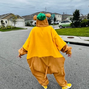 Puede incluir: Una persona con un disfraz de cuerpo entero que se asemeja a un personaje de dibujos animados. El disfraz es principalmente amarillo y marr&oacute;n, con un sombrero verde y zapatos amarillos. La persona est&aacute; de pie en una carretera pavimentada en una zona residencial.