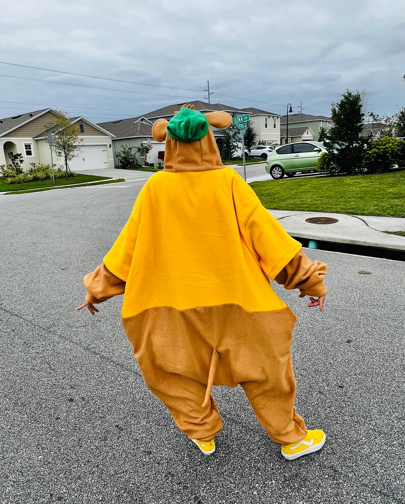 Puede incluir: Una persona con un disfraz de cuerpo entero que se asemeja a un personaje de dibujos animados. El disfraz es principalmente amarillo y marr&oacute;n, con un sombrero verde y zapatos amarillos. La persona est&aacute; de pie en una carretera pavimentada en una zona residencial.