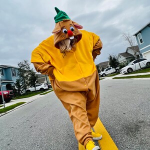 Puede incluir: Una persona con un disfraz de conejo amarillo y marr&oacute;n con un gorro verde y zapatillas amarillas. El disfraz tiene una capucha con cara de conejo con una nariz roja y orejas largas. La persona camina por una carretera.