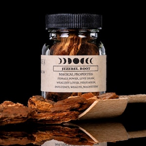 Op de afbeelding: Een doorzichtige glazen pot met een zwarte deksel gevuld met bruine Jezebel-wortelstukjes. Het etiket toont maanfasen en de tekst: JEZEBEL ROOT, MAGICAL PROPERTIES: FEMALE POWER, LOVE DRAW, WEALTHY LOVER, PERSUASION, INFLUENCE, WEALTH, MAGNETISM. Een houten schepje ligt ernaast.