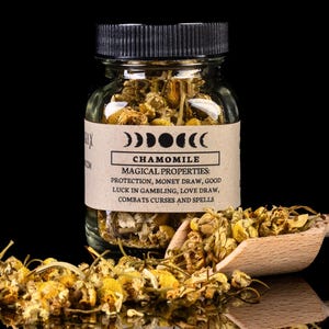 May include: A clear glass jar filled with dried chamomile flowers, labeled with "Chamomile" and its magical properties. A wooden scoop holds more chamomile, with loose flowers scattered around. The background is black.
