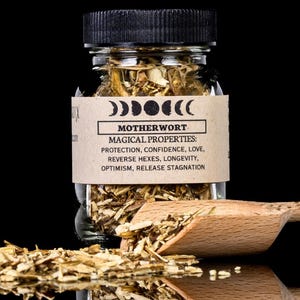 Pode incluir: Um frasco de vidro transparente cheio de ervas secas, rotulado "MOTHERWORT" com "PROPRIEDADES MÁGICAS" listadas. Uma colher de madeira repousa ao lado do frasco, e ervas soltas estão espalhadas.