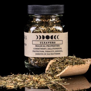May include: A small glass jar filled with dried herbs, labeled "CLEAVERS" with "MAGICAL PROPERTIES" listed below. A wooden scoop holds more herbs, with a scattering of herbs in front. The background is black.