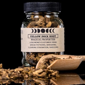 Pode incluir: Um frasco de vidro cheio de raiz de doca amarela, com tampa preta e um rótulo que diz "YELLOW DOCK ROOT". Uma colher de madeira e pedaços soltos de raiz estão em frente ao frasco. O rótulo lista propriedades mágicas.