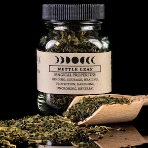 May include: A glass jar filled with dried green nettle leaf. The jar has a black lid and a brown label with the text '00000(( Nettle Leaf Magical Properties: Binding, Courage, Healing, Protection, Banishing, Uncrossing, Reversal'.