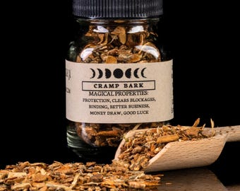 May include: A glass jar filled with brown bark, labeled "CRAMP BARK" with the text "MAGICAL PROPERTIES: PROTECTION, CLEARS BLOCKAGES, BINDING, BETTER BUSINESS, MONEY DRAW, GOOD LUCK".