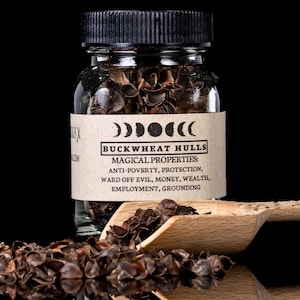 Peut inclure: Un bocal en verre transparent rempli de balles de sarrasin, étiqueté "BUCKWHEAT HULLS" et "MAGICAL PROPERTIES". Une cuillère en bois et des balles éparpillées sont devant le bocal. L'étiquette énumère des propriétés telles que l'anti-pauvreté et la protection.