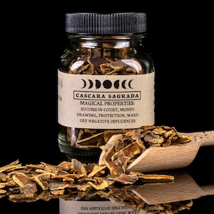 Puede incluir: Un frasco de vidrio lleno de corteza de Cascara Sagrada seca. El frasco tiene una etiqueta marrón con el texto "Cascara Sagrada" y "Propiedades mágicas: Éxito en la corte, dinero, dibujo, protección, protección contra influencias negativas".