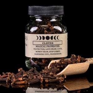 May include: A glass jar filled with brown cloves, with a black label that reads "Cloves" and lists magical properties: protection, love draw, luck, money draw, stop gossip, banishing evil, friendships.