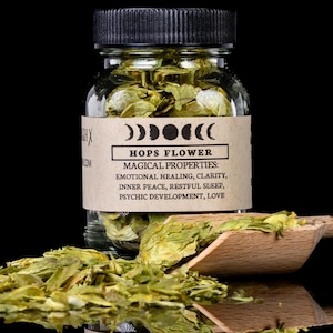 Puede incluir: Un frasco de vidrio transparente lleno de flores de lúpulo secas, con una tapa negra y una etiqueta que dice "HOPS FLOWER MAGICAL PROPERTIES: EMOTIONAL HEALING, CLARITY, INNER PEACE, RESTFUL SLEEP, PSYCHIC DEVELOPMENT, LOVE". Se ve parcialmente una cuchara de madera.