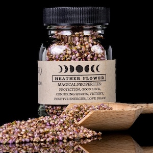 Puede incluir: Un frasco de vidrio transparente lleno de flores de brezo, una cuchara de madera y flores dispersas. El frasco tiene una tapa negra y una etiqueta con el texto "HEATHER FLOWER" y "MAGICAL PROPERTIES: PROTECTION, GOOD LUCK, CONJURING SPIRITS, VICTORY, POSITIVE ENERGIES, LOVE DRAW."