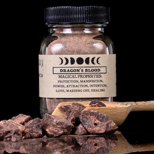 May include: A glass jar of Dragon's Blood powder with a black lid, a wooden scoop, and pieces of resin. The label lists magical properties: protection, manifestation, power, attraction, intention, love, warding off, and healing.