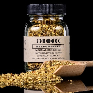 Puede incluir: Un frasco de vidrio transparente con tapa negra lleno de hierbas de reina de los prados secas de color dorado. Una cuchara de madera contiene más hierbas, con algunas derramándose. La etiqueta dice "MEADOWSWEET" y enumera propiedades mágicas.
