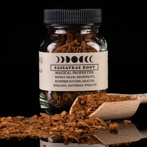 Pode incluir: Um frasco de vidro cheio de casca de raiz de sassafras marrom. O frasco tem uma tampa preta e um rótulo marrom que diz "Raiz de Sassafras" e lista suas propriedades mágicas: "Atrair dinheiro, prosperidade, sucesso nos negócios, saúde, cura, riqueza material".