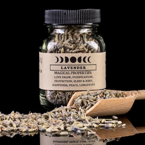Puede incluir: Un frasco de vidrio lleno de flores de lavanda secas. El frasco tiene una tapa negra y una etiqueta marrón que dice "LAVANDA" y enumera las propiedades mágicas de la lavanda: atraer el amor, purificar, proteger, dormir y descansar, felicidad, paz, longevidad.