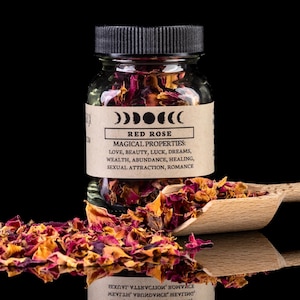 Puede incluir: Un frasco de vidrio lleno de pétalos de rosa roja, etiquetado como "RED ROSE" con "MAGICAL PROPERTIES" enumeradas a continuación. Una cuchara de madera contiene más pétalos. El fondo es negro, con una superficie espejada.