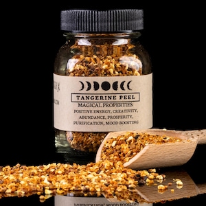 Puede incluir: Un frasco de vidrio transparente lleno de cáscara de mandarina seca, con tapa negra. Una cuchara de madera contiene más cáscara, y algo está esparcido en la superficie. La etiqueta dice "Tangerine Peel" y enumera propiedades mágicas.
