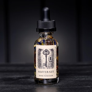 May include: A clear glass bottle with a black dropper top, filled with a liquid and botanicals. A label on the bottle reads "MASTER KEY DIVINE PERMISSION" with a key illustration. The bottle is on a dark surface.