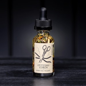 May include: A clear glass bottle with a black dropper top, filled with a golden liquid and botanicals. A label features scissors cutting cords, with the text "CUTTING CORDS / SEVER TIES / RELEASE". The bottle is on a dark surface.