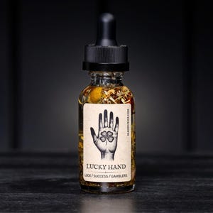 Pode incluir: Uma garrafa de vidro com conta-gotas preto, cheia de líquido transparente e botânicos. O rótulo diz "LUCKY HAND" com uma ilustração de mão e trevo de quatro folhas. O texto "LUCK / SUCCESS / GAMBLERS" está abaixo.