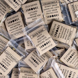 May include: An assortment of small, clear plastic bags filled with dried herbs. Each bag has a tan paper label with black text, including the words "Rock Rose," "Chamomile," and "Lady's Mantle." The labels also feature moon phase graphics.