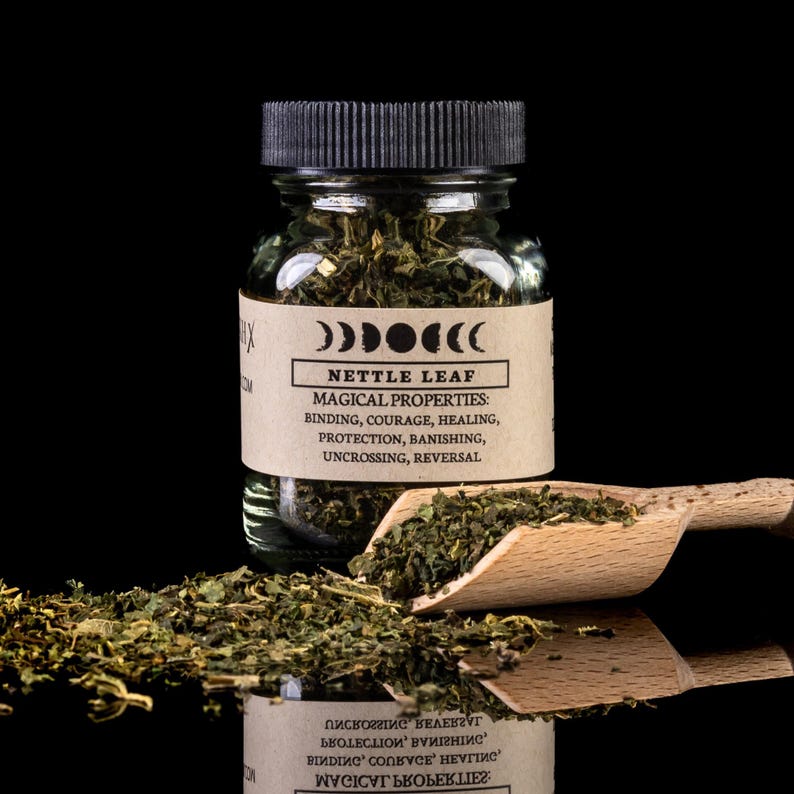 May include: A glass jar filled with dried green nettle leaf. The jar has a black lid and a brown label with the text '00000(( Nettle Leaf Magical Properties: Binding, Courage, Healing, Protection, Banishing, Uncrossing, Reversal'.