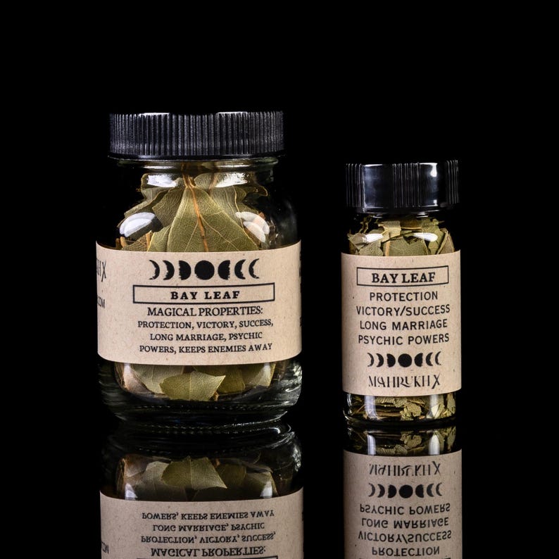 May include: Two glass jars filled with dried bay leaves. The jars have brown paper labels with the text "Bay Leaf" and a list of magical properties, including protection, victory, success, long marriage, psychic powers, and keeps enemies away. The labels also feature a crescent moon symbol.