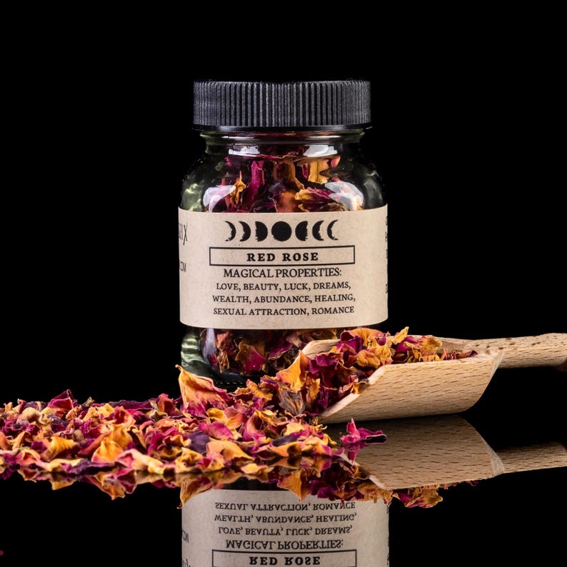 May include: A glass jar filled with red rose petals, labelled "RED ROSE" with "MAGICAL PROPERTIES" listed below. A wooden scoop holds more petals. The background is black, with a mirrored surface.