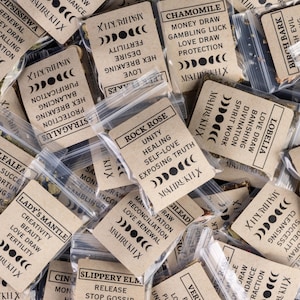 May include: A collection of small plastic bags filled with dried herbs, each with a tan paper label. The labels feature text such as "Rock Rose" and "Chamomile", moon phase symbols, and the brand name "MAHRUKH X".