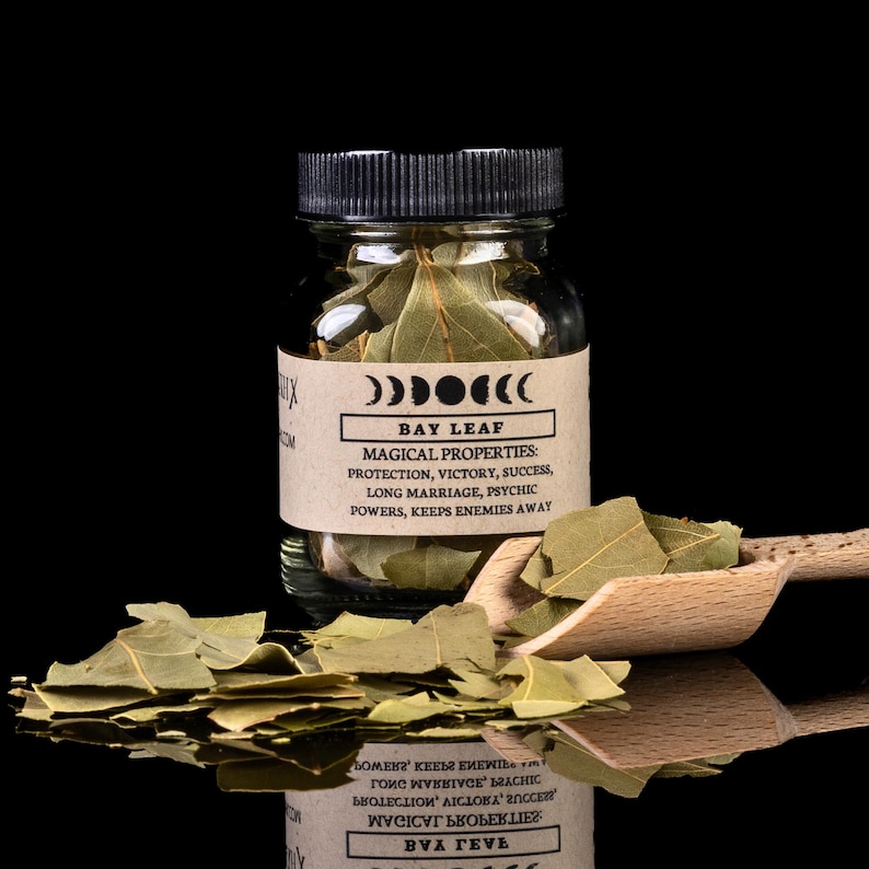 May include: A glass jar filled with dried bay leaves and a wooden scoop with more bay leaves. The jar has a black label with white text that reads 'BAY LEAF' and 'MAGICAL PROPERTIES: PROTECTION, VICTORY, SUCCESS, LONG MARRIAGE, PSYCHIC POWERS, KEEPS ENEMIES AWAY'.