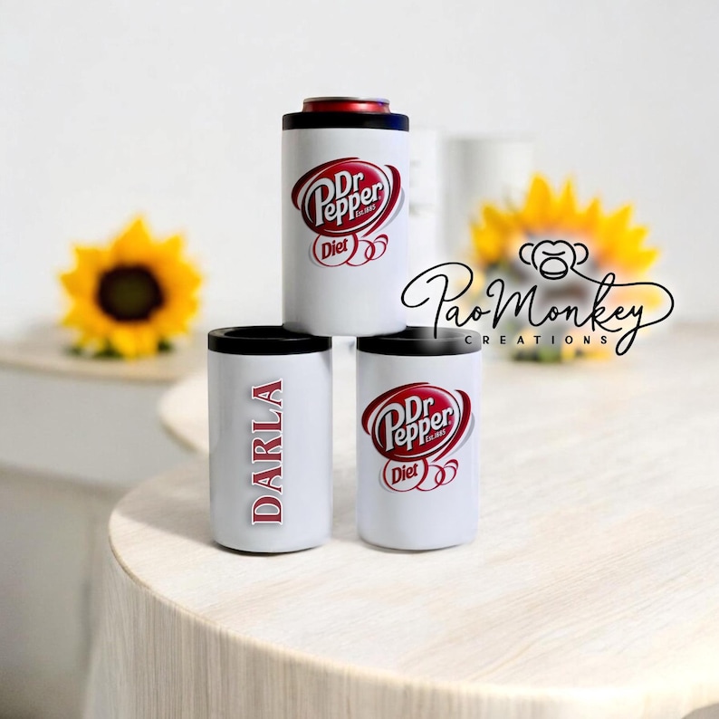 May include: Three white can coolers, one with the text "DARLA" in red, and two with the Diet Dr Pepper logo. The coolers are on a white table.
