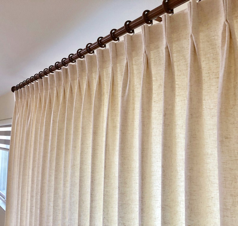 May include: Close-up of a cream-colored curtain with pleats hanging from a wooden rod.