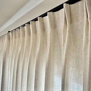 May include: Off-white, textured fabric curtains featuring a pinch pleat header design. The curtains are suspended from a thin, black horizontal rod mounted near the ceiling, creating uniform vertical folds that extend downward along the fabric.