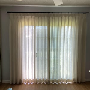 May include: A pair of white sheer curtains with pleats hang from a black curtain rod in front of a sliding glass door. The curtains are drawn and the room is lit by natural light.