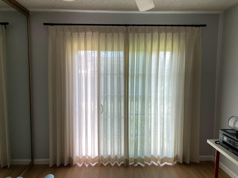 May include: A pair of white sheer curtains with pleats hang from a black curtain rod in front of a sliding glass door. The curtains are drawn and the room is lit by natural light.