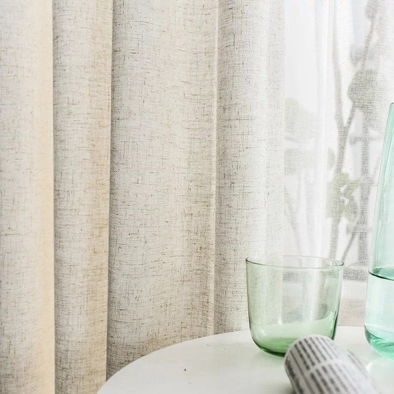 May include: Two sheer linen curtains hang in a window, with a clear glass tumbler and a glass bottle on a white table in front of them.