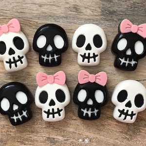 May include: A collection of eight skull-shaped embellishments. The skulls are in black and white, some with pink bows. The skulls have large eye sockets, a nose cavity, and a stitched mouth. The embellishments are arranged on a wooden surface.
