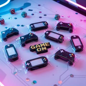May include: An overhead shot of various gaming-themed candies, including game controllers, handheld consoles, and dice. The candies are black with colorful accents and are individually wrapped. The phrase "GAME ON" is spelled out in yellow and black candy.
