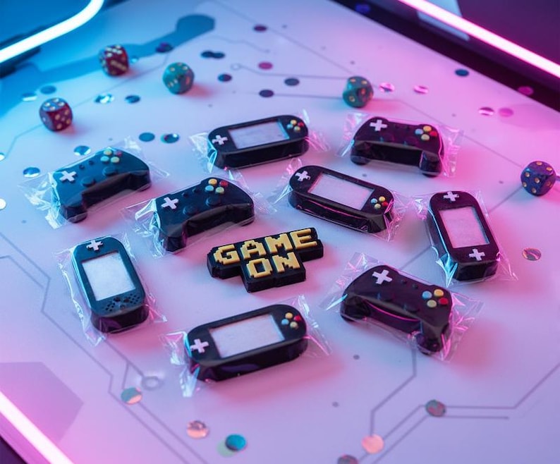 May include: An overhead shot of various gaming-themed candies, including game controllers, handheld consoles, and dice. The candies are black with colorful accents and are individually wrapped. The phrase "GAME ON" is spelled out in yellow and black candy.