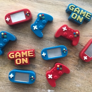 May include: A collection of colorful gaming-themed accessories. The image features red and blue game controllers, handheld consoles, and the phrase "GAME ON" in pixelated lettering. The accessories have white, yellow, and green accents.