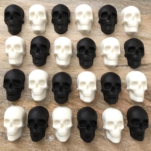 May include: An overhead shot displays a collection of small skull figurines. The skulls alternate between black and white, arranged in a grid pattern. The skulls are made of a solid material and have detailed facial features.