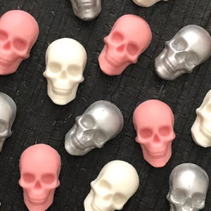 May include: A collection of small skull-shaped objects in matte pink, matte white, and metallic silver finishes. The skulls are arranged in a scattered pattern, each featuring detailed eye sockets, nasal cavities, and teeth, presented in a uniform size.