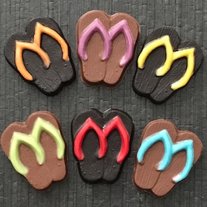 May include: Six pairs of chocolate flip-flop sandals with different colored straps. The straps are orange, purple, yellow, green, red, and blue.
