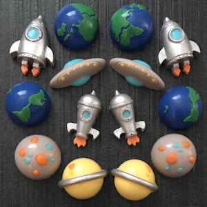 May include: A collection of space-themed embellishments, including silver rockets with orange flames, blue and green Earths, beige UFOs, and planets with rings and colorful spots. These decorative elements are ideal for crafting projects.
